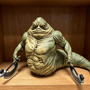 May include: A green, slug-like creature figurine with a muscular torso, wrinkled skin, and a tail-like lower body. The figure has a stern facial expression and holds a curved, metallic-looking blade in each hand, extending outward to the sides.