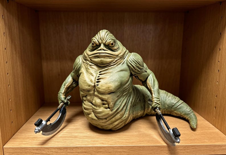May include: A green, slug-like creature figurine with a muscular torso, wrinkled skin, and a tail-like lower body. The figure has a stern facial expression and holds a curved, metallic-looking blade in each hand, extending outward to the sides.