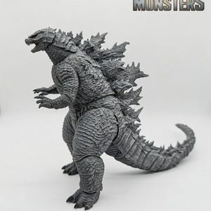 May include: A grey Godzilla action figure with a textured, scaly surface, sharp dorsal fins, and a long tail. The figure is positioned in profile, and the text "KING OF THE MONSTERS" with a gold crown icon is visible in the upper right corner.
