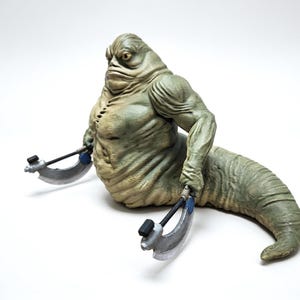 May include: A green, slug-like creature figurine with a muscular upper body and a long, tapering tail. The figure has a wrinkled, textured skin surface and is holding two silver-colored, crescent-shaped bladed weapons in its hands.