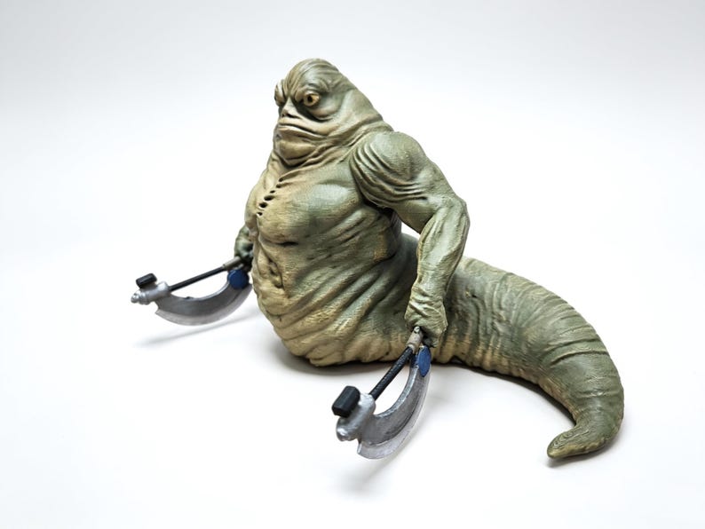 May include: A green, slug-like creature figurine with a muscular upper body and a long, tapering tail. The figure has a wrinkled, textured skin surface and is holding two silver-colored, crescent-shaped bladed weapons in its hands.