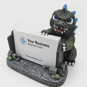 May include: A dark green monster figurine with blue dorsal fins holding a stack of white business cards. The base is textured like gray stone rubble. The card displays a blue G logo, the text Your Business Business Card, and a website URL.