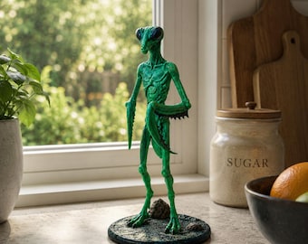 May include: A green praying mantis figurine with detailed texture stands on a round, textured base. The insect-like sculpture has large eyes and is posed with its arms bent. The figurine is displayed on a countertop near a window and a sugar jar.