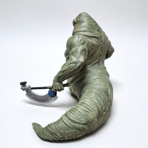 May include: A green, muscular, slug-like creature figurine with a textured, ridged body and a hunched back. The figure is holding a small, silver-colored curved blade weapon with a dark handle and a blue circular accent on the side.