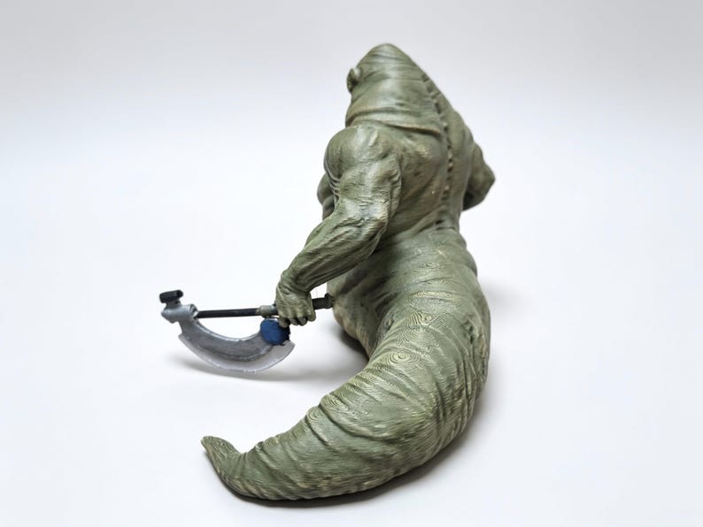 May include: A green, muscular, slug-like creature figurine with a textured, ridged body and a hunched back. The figure is holding a small, silver-colored curved blade weapon with a dark handle and a blue circular accent on the side.