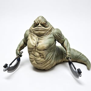 May include: A pale green, slug-like creature figurine with a muscular torso, heavy facial features, and a long, tapering tail. The figure holds a curved, silver-colored blade weapon in each hand, featuring black accents on the handles and blades.