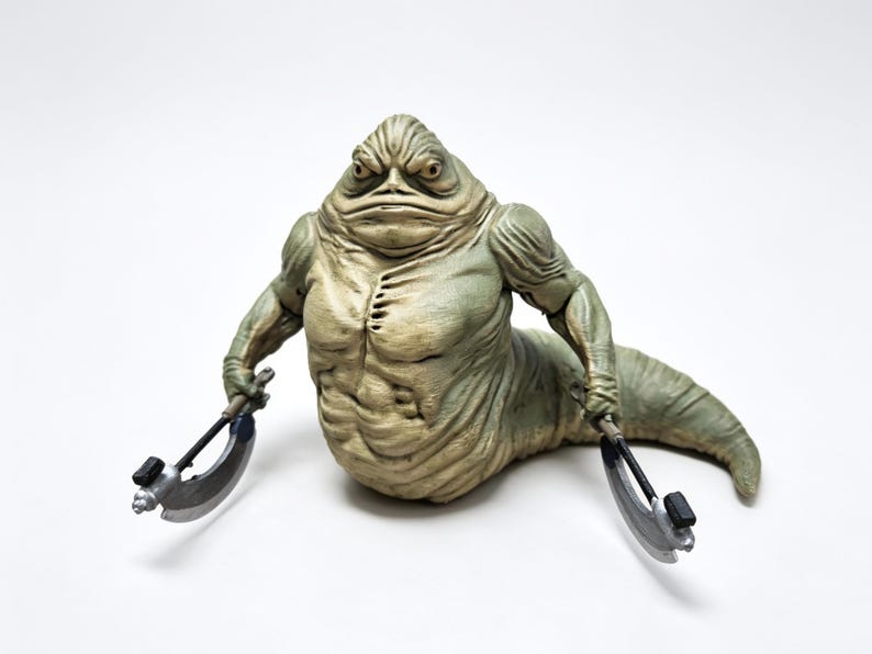 May include: A pale green, slug-like creature figurine with a muscular torso, heavy facial features, and a long, tapering tail. The figure holds a curved, silver-colored blade weapon in each hand, featuring black accents on the handles and blades.