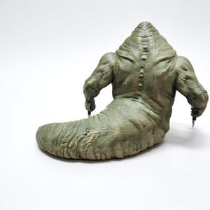 May include: A green, slug-like creature figurine with a muscular upper body, a thick tail, and a row of small spikes running down its back. The figure has two arms extending outward, each holding a small, sharp, metallic-colored pointed object.