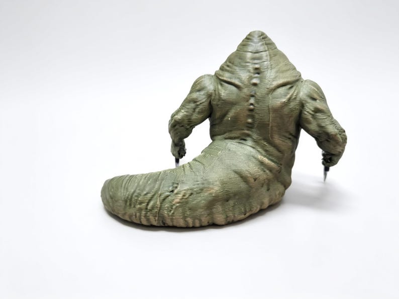May include: A green, slug-like creature figurine with a muscular upper body, a thick tail, and a row of small spikes running down its back. The figure has two arms extending outward, each holding a small, sharp, metallic-colored pointed object.
