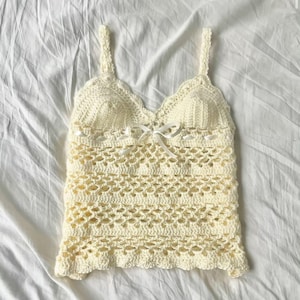 May include: A cream-colored crochet camisole featuring a V-neckline with a scalloped edge, thin shoulder straps, and a delicate white ribbon bow centered at the bust. The garment is constructed with an openwork stitch pattern throughout the body.