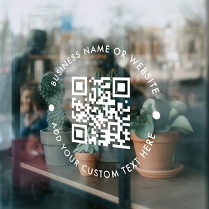 May include: A white circular decal applied to a glass surface featuring a central QR code. The text "BUSINESS NAME OR WEBSITE" is printed along the top arc, and "ADD YOUR CUSTOM TEXT HERE" is printed along the bottom arc of the white circle.
