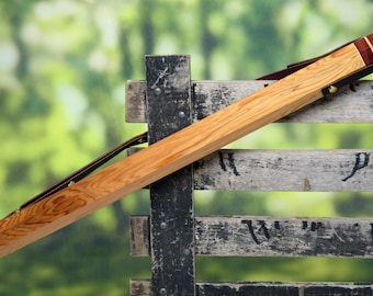 May include: A wooden case with a dark brown leather strap and accents. The case is made of light-colored wood with a reddish-brown end cap. The case is resting on a weathered wooden fence.