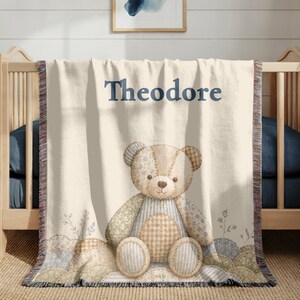 May include: A cream-colored woven throw blanket featuring the name Theodore in dark blue text above a central illustration of a patchwork teddy bear. The bear is composed of various tan, blue, and white patterned fabrics with stitched details.