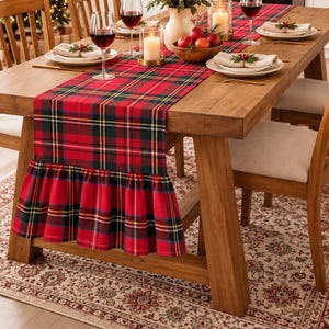 May include: A rectangular table runner featuring a classic red, green, and yellow plaid pattern with a gathered ruffle edge. The fabric runner is draped across a wooden dining table, showcasing a festive textile design with clean, straight lines.