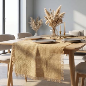 May include: A rectangular tan hessian table runner featuring a textured woven fabric and frayed edges on all sides. The runner is draped over a wooden dining table, providing a neutral base for table settings and decorative centrepieces.