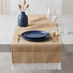 Könnte beinhalten: Ein beigefarbener Tischläufer aus Jute mit weißer Fransenborte, dazu eine marineblaue Keramikvase mit getrockneten Mohnkapseln, ein marineblaues Tellerset, goldfarbenes Besteck, ein Weinglas aus Kristall und ein Kristallglas auf einer grauen Fläche.