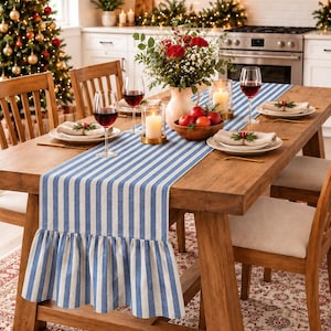 May include: A blue and white striped table runner with a ruffled edge draped over a wooden dining table. The runner features vertical stripes and a gathered fabric border at the ends, providing a decorative accent for the table setting.