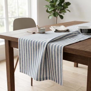 May include: A rectangular table runner featuring a pattern of vertical blue and white stripes. The fabric is draped over a wooden table, with a small ceramic bowl, a folded beige napkin, and a black and white patterned bowl placed on top.