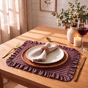 May include: A rectangular dark maroon placemat with white pinstripes and a ruffled border, featuring a round wooden charger plate, a white ceramic dinner plate, a beige linen napkin held by a woven rattan napkin ring, and gold cutlery.