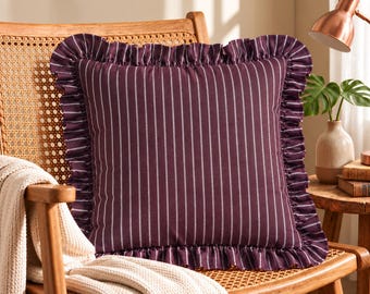 May include: A square throw pillow in a deep plum purple color featuring thin vertical white stripes. The pillow is finished with a ruffled fabric border along all four edges, creating a decorative frame around the striped center panel.