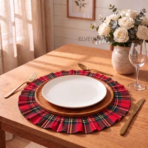 May include: A round fabric placemat featuring a red, green, and yellow plaid pattern with a ruffled edge. The placemat is topped with a circular wooden charger plate and a plain white ceramic dinner plate. A gold fork and knife are placed nearby.