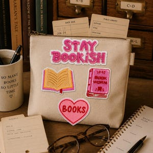 Może przedstawiać: Beżowy piórnik z płótna zapinany na zamek, ozdobiony haftowanymi naszywkami, w tym napisem STAY BOOKISH, otwartą książką, różową książką z napisem BOOK WORM oraz różowym sercem ze słowem BOOKS. Piórnik posiada metalowy zamek błyskawiczny i boczną zawieszkę.