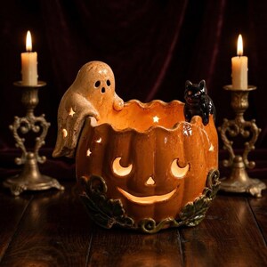 Puede incluir: Un cuenco de cerámica naranja con forma de calabaza, cara tallada, una figura de fantasma beige en el borde y un pequeño gato negro. La calabaza tiene recortes en forma de estrella y hojas verdes en la base, iluminada desde el interior.