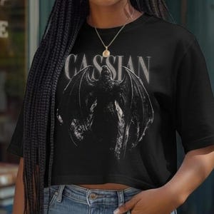 May include: A black short-sleeved t-shirt featuring a graphic print of a winged creature and the text CASSIAN in white lettering. The shirt is paired with blue denim jeans and a gold-toned necklace with a small circular pendant.