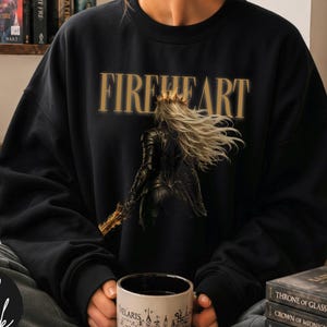 May include: A black crewneck sweatshirt featuring a graphic print of a figure in dark armour with long flowing hair and a crown, positioned below the gold text FIREHEART. The design is centred on the chest of the long-sleeved garment.
