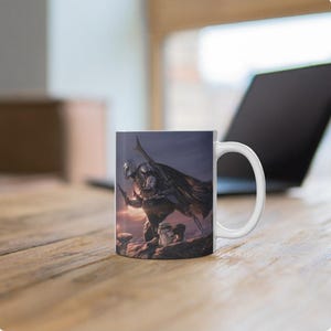 May include: A white ceramic mug featuring a printed illustration of a silver-armored warrior in a cape kneeling on a rocky surface with a small green creature. The warrior holds a blaster and has a long rifle strapped to their back.