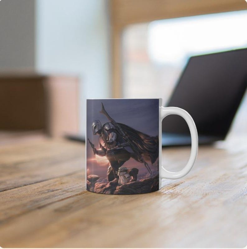 May include: A white ceramic mug featuring a printed illustration of a silver-armored warrior in a cape kneeling on a rocky surface with a small green creature. The warrior holds a blaster and has a long rifle strapped to their back.