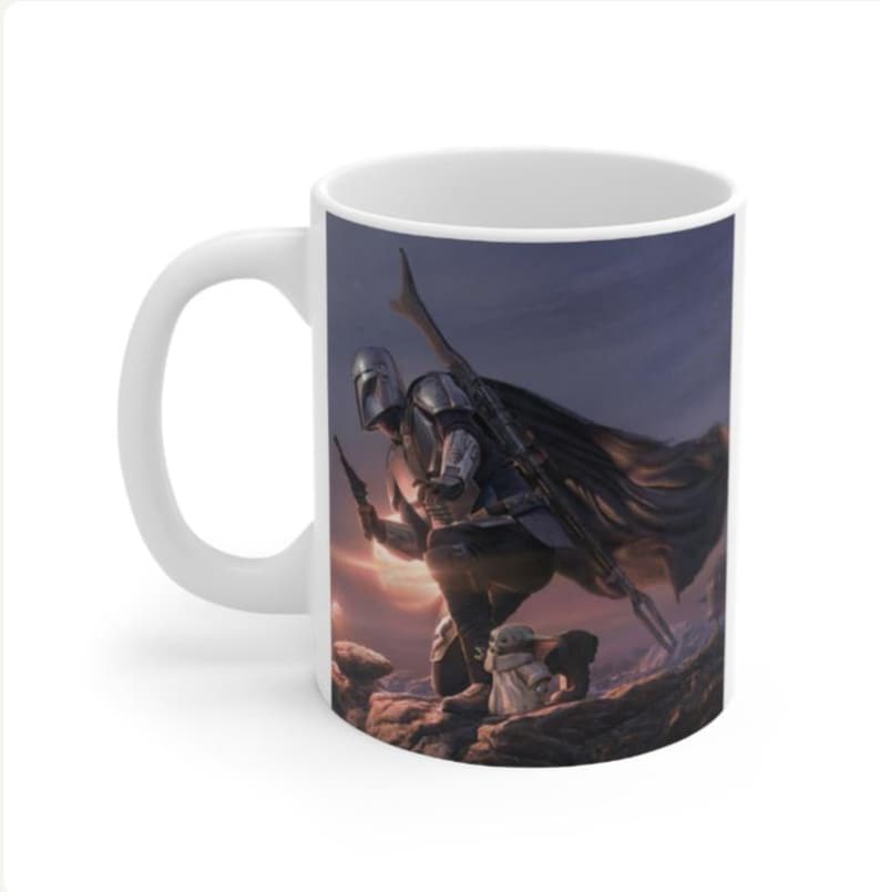 May include: A white ceramic mug featuring a printed illustration of a silver-armored warrior kneeling on rocky terrain, holding a blaster and a long staff, with a small green creature in a tan robe standing beside them under a dark sky.