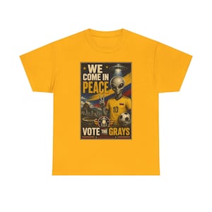 May include: Mustard yellow t-shirt featuring a graphic of an alien in a soccer jersey holding a soccer ball. The graphic includes the text "WE COME IN PEACE" and "VOTE THE GRAYS".