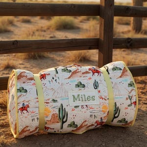 May include: Rolled-up, patterned blanket with a yellow strap and trim. The blanket features a desert landscape design with cacti, teepees, and cowboys. The name "Miles" is embroidered on the blanket.