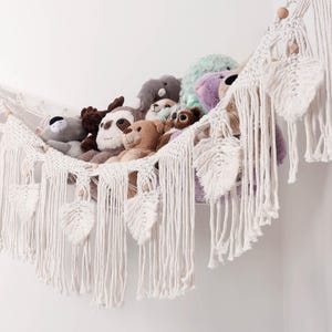 May include: A macrame wall hanging in white, featuring a hammock design filled with various plush toys. The macrame has a fringe and leaf-shaped details. The plush toys include a sloth, bear, and other stuffed animals.