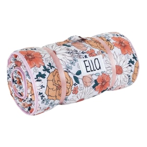 May include: Rolled-up floral picnic blanket with a white background and orange, peach, and teal flower pattern. The blanket is secured with pink straps and features the word "ELLO" on a white label.