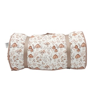May include: Rolled-up, quilted mat with a mushroom and foliage pattern. The mat is primarily white with brown and tan illustrations. It is secured with a tan strap and has a tan border. The mat is designed for outdoor use.