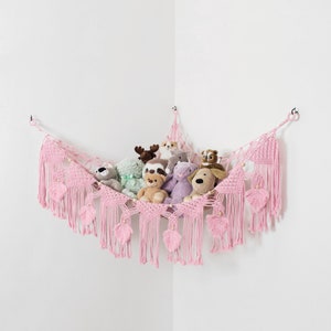 May include: A pink macrame corner hammock for stuffed animals, featuring intricate knotted patterns, hanging fringe, and decorative leaf-shaped accents. The net is suspended by rings and holds a variety of plush toys in a corner arrangement.