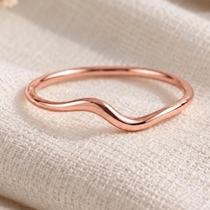 May include: A thin rose gold ring featuring a smooth, polished finish and a subtle wave-like curve along the band. The metal has a warm, metallic lustre, and the design is continuous, forming a simple, elegant, and fluid shape for the finger.