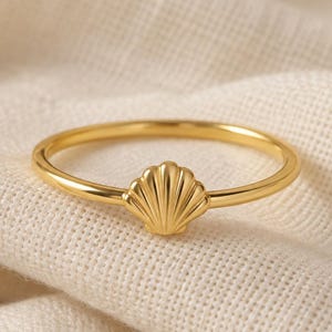 May include: A thin gold ring featuring a small, centred seashell charm. The band is smooth and circular, while the seashell element displays vertical ridges and a fan-like shape. The metal has a polished, reflective finish throughout the entire piece.