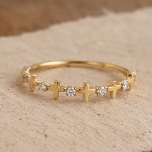 May include: A thin gold ring featuring an alternating pattern of small, polished gold crosses and clear, round-cut stones set along the band. The design is symmetrical, with the crosses and stones spaced evenly around the circumference of the ring.
