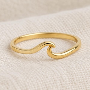 May include: A polished gold ring featuring a continuous band that curves upwards into a single, smooth wave shape. The metal has a reflective, high-shine finish, and the design is simple, consisting of a thin, rounded band that forms the wave motif.