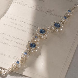 May include: A delicate silver bracelet with blue gemstone accents and faux pearls. The bracelet is displayed on a wedding invitation with the text "THE WEDDING OF Jennifer Lynn Powers and Robert Allen Everson". The invitation is cream-colored.