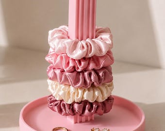 May include: A pink vertical stand with a circular base holds five stacked satin scrunchies in shades of light pink, rose, mauve, cream, and dark berry. A thin gold-toned ring and a pair of small stud earrings rest on the base of the stand.