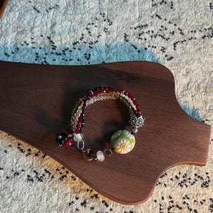 Könnte beinhalten: Ein mehrsträngiges Armband mit roten, olivgrünen und klaren facettierten Perlen. Es enthält eine runde, lindgrüne Keramikperle mit weißem Gänseblümchenmuster, eine verzierte silberfarbene Perlenkappe und einen kupferfarbenen herzförmigen Karabinerverschluss.