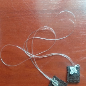 May include: Two brown scapulars with silver pendants on clear cords. One scapular has a silver cross pendant, and the other has a silver pendant with a religious figure. The scapulars are on a brown wooden surface.