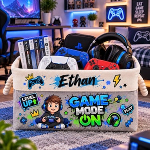 Puede incluir: Una cesta de almacenamiento rectangular de tela con el nombre Ethan, un personaje de dibujos animados con auriculares y el texto Game Mode On y Level Up. La cesta contiene mandos de videojuegos, auriculares y cajas de juegos.