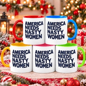May include: Five white ceramic mugs with dark blue text that reads AMERICA NEEDS NASTY WOMEN. Each mug features a different colored handle and interior rim, including red, blue, yellow, pink, and green, arranged in a two-row display.