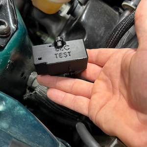May include: A black rectangular device with the text "EEC TEST" printed on it. The device is held in front of a car engine bay. The engine bay has green and black components.