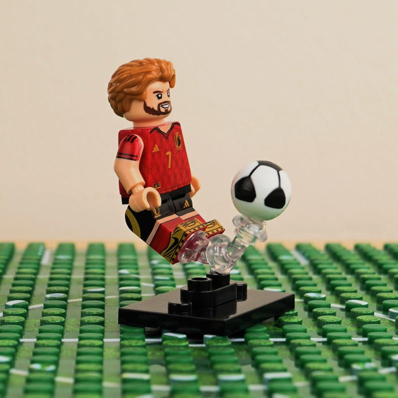 May include: A Lego figurine of a soccer player in a red jersey with the number 7, kicking a black and white soccer ball. The figurine is on a black base, set against a green field background.